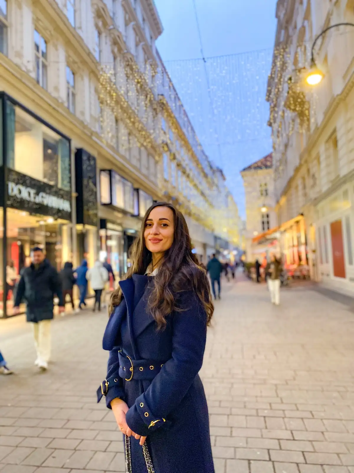 A portrait of a smiling woman with long dark hair wearing a navy blue coat. She is standing in the middle of a paved pedestrian shopping street (The Graben) under elaborate hanging Christmas lights that look like chandeliers. Luxury shop fronts are visible in the background.