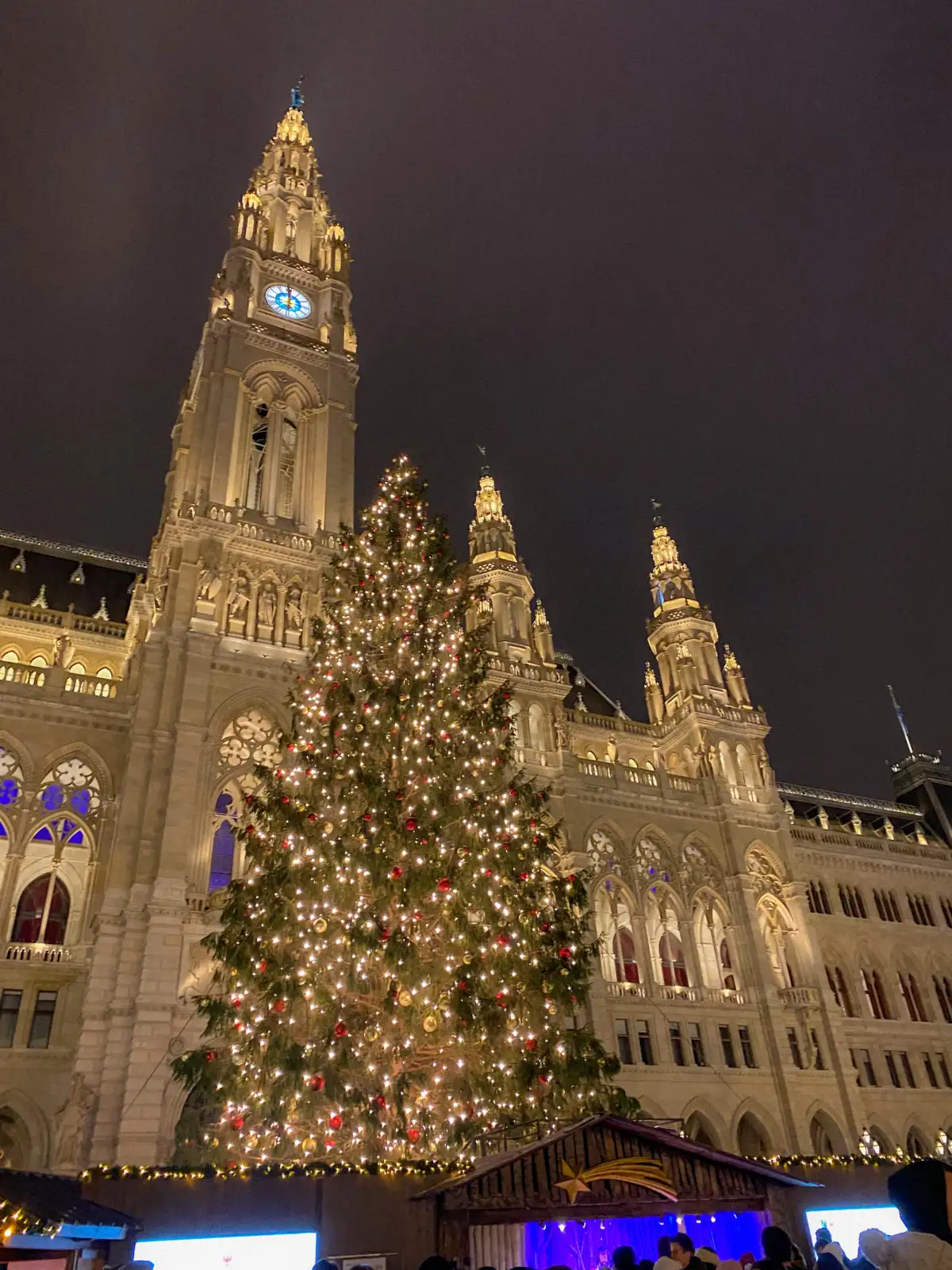 A night view of the Vienna City Hall (Rathaus) tower illuminated in warm light. In the foreground stands a massive Christmas tree covered in lights and red ornaments, towering over the roofs of Christmas market stalls.